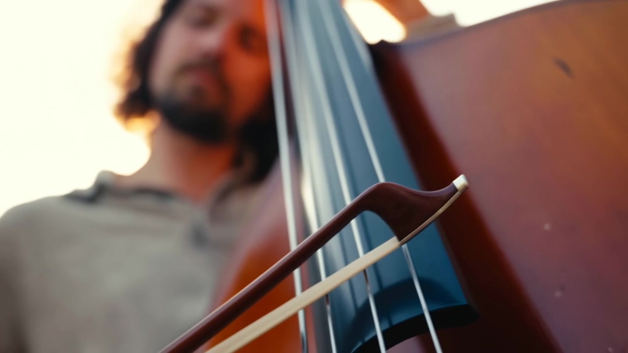 Extreme close view of upright double bass being bowed, with bow hair pressing across metal strings over dark fingerboard and glossy reddish brown body, creating warm concert mood and rich tactile
