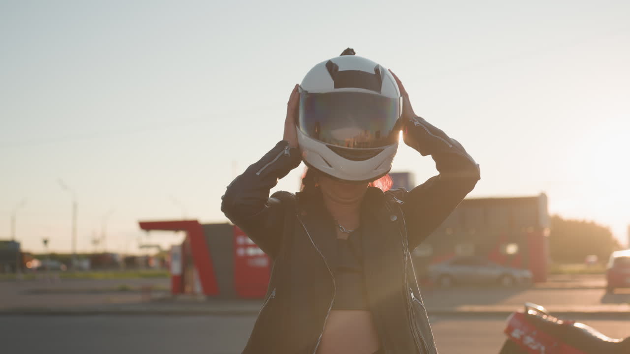 Female biker prepares to put on helmet wearing leather jacket standing in urban setting as another rider drops off motorcycle with partial view of companion nearby under warm sunlight