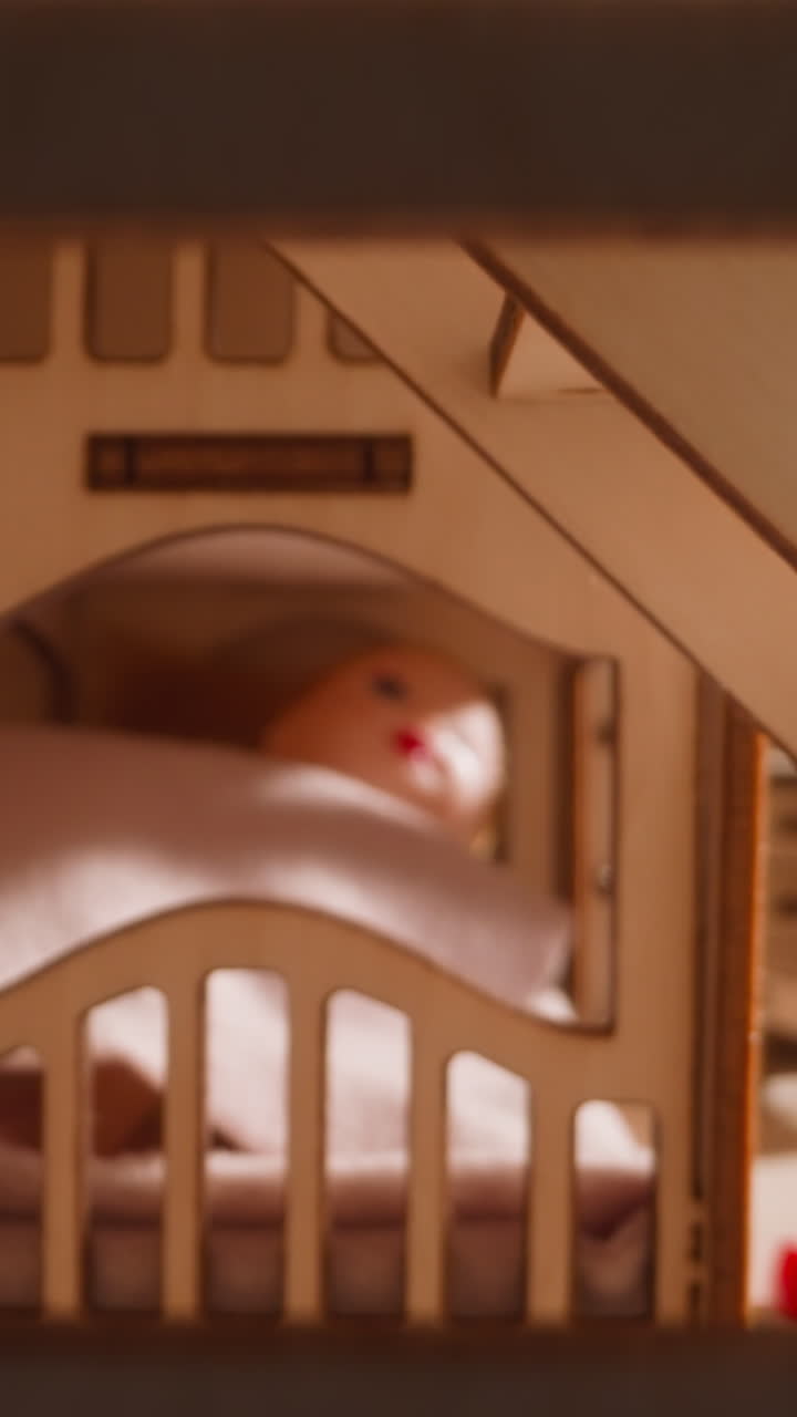 La mano di un bambino piccolo calma una bambola coperta con un plaid rosa nel letto di una casa giocattolo di legno, vista ravvicinata con zoom indietro che mostra la stanza con gradini e finestra.