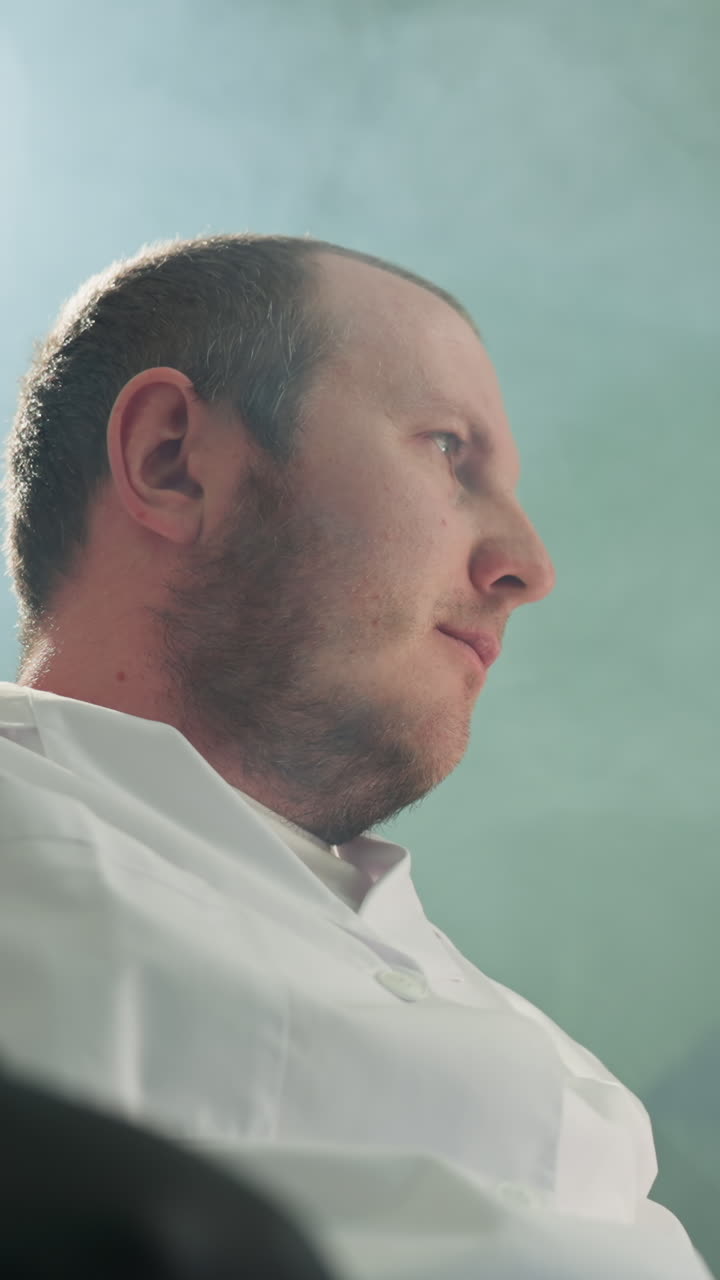 крупный вид ученого в белом лабораторном халате, сидящего и отдыхающего с закрытыми глазами, рукой на лбу, в лабораторной обстановке, с полками и освещенным микроскопом и отражением света