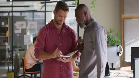 Happy-diverse-business-people-holding-notebook-and-discussing-work-at-office