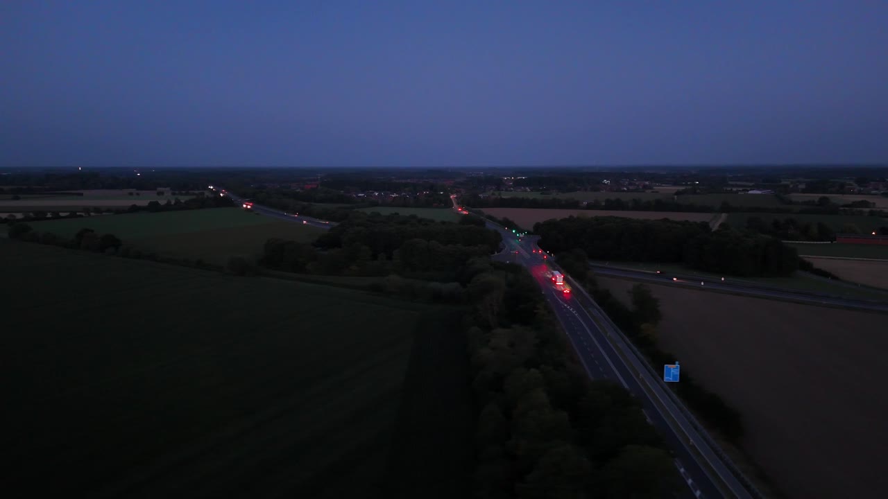 Illuminated expressway between rural farm fields at dusk. American countryside with driving cars and headlights at night. Aerial panorama wide shot