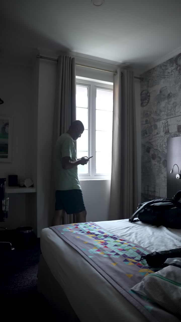 asiático del sur maduro en ropa casual usando teléfono inteligente cerca de la ventana en una habitación de hotel moderna, disfrutando de la luz natural y la conectividad durante un viaje de negocios. video vertical