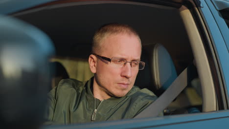 close-up front view of a man wearing glasses and a green jacket sitting in the driver s seat of a car. he is looking straight and gazing out the window, appearing focused and contemplative