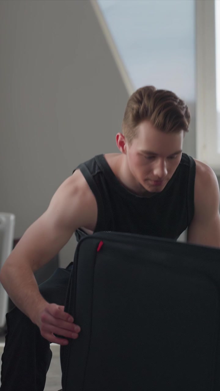 Young man in black sleeveless shirt sits on chair handling. Man adjusts his position while seated indoors. Scene features gray walls and angled window suggesting home workout