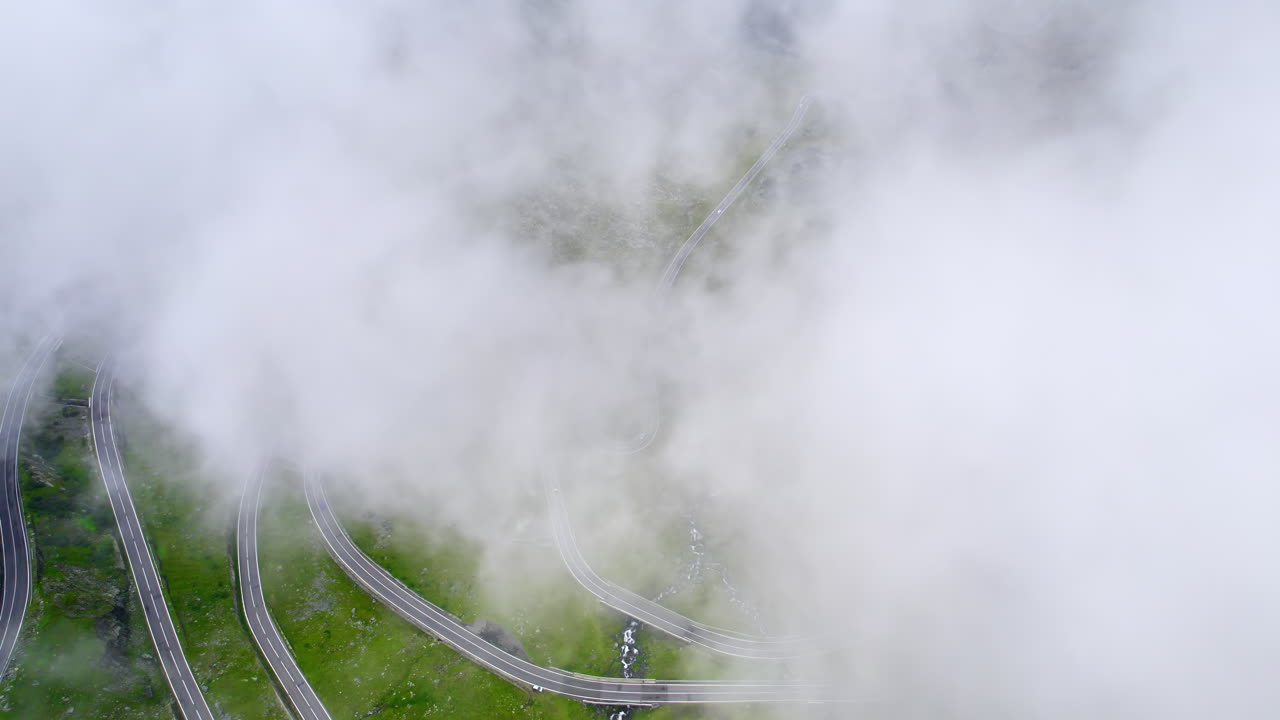 Aerial drone view of nature in Romania. Transfagarasan route in Carpathian mountains with moving cars visible through clouds, rocky slopes with greenery