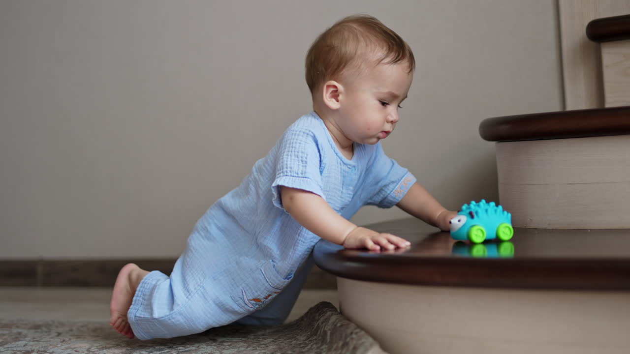 Peaceful Caucasian baby boy in blue romper is beside the staircase. A toy lies on the stair but the kid looks aside.
