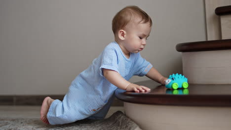 peaceful caucasian baby boy in blue romper is beside the staircase. a toy lies on the stair but the kid looks aside.