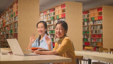 asian woman students with headphones smiling to camera and waving hands while reading books on the table with laptop in the library