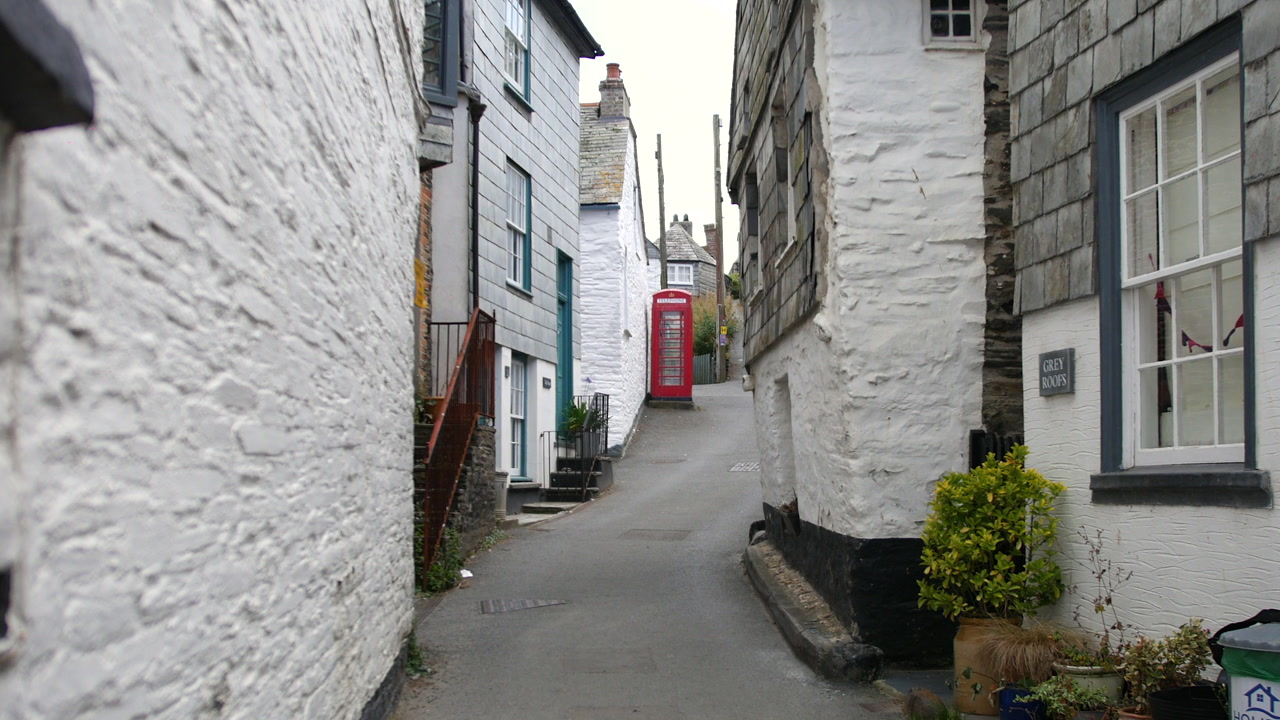 A street view in Port Isaac, England, showing a traditional red telephone box along a winding lane lined with historic cottages and slate-clad homes in the fictional village of Portwenn