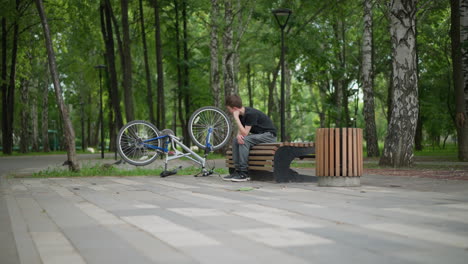 white boy sits thoughtfully on park bench, his hand resting under his chin, beside him is an upside-down bicycle, and in the background, slightly blurred pigeons are walking on the park's floor