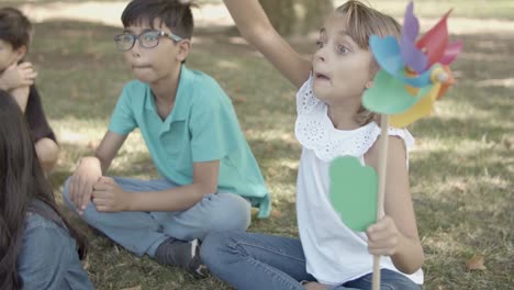 Süßes-Kaukasisches-Mädchen,-Das-Papierfächer-Hält,-Fußballball-Fängt-Und-Ihn-Wegwirft,-Während-Er-Mit-Freunden-Auf-Gras-Im-Park-Sitzt