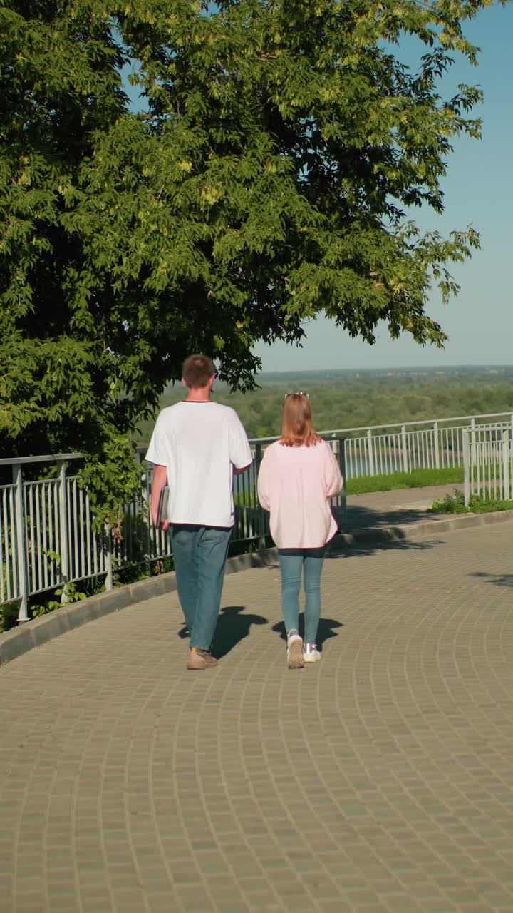 niña en camiseta rosa y vaqueros caminando cerca con su amiga sosteniendo una computadora portátil en la mano izquierda a lo largo de un carril de hierro con un fondo pintoresco con un vasto bosque, un río que fluye y un cielo despejado