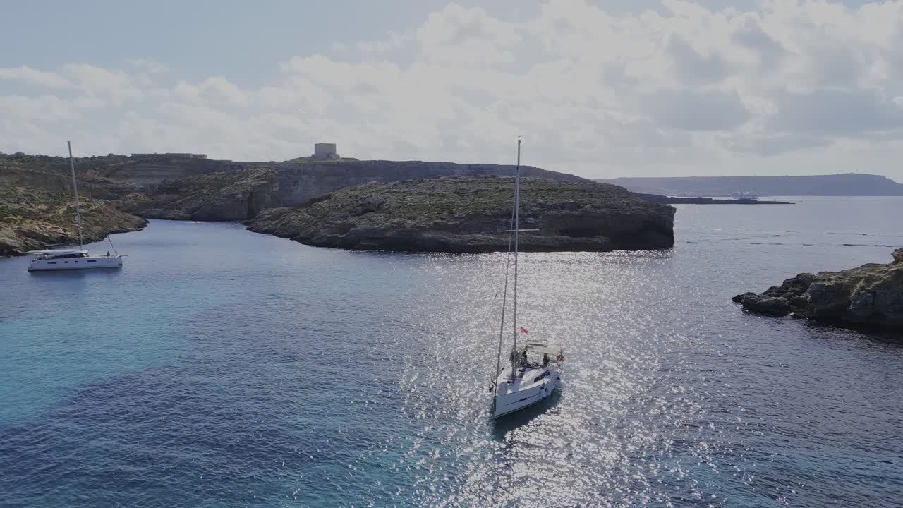 A calm rotating drone shot shows two anchored yachts at Blue Lagoon, with sunlight reflecting on the water and the Comino coastline in the background