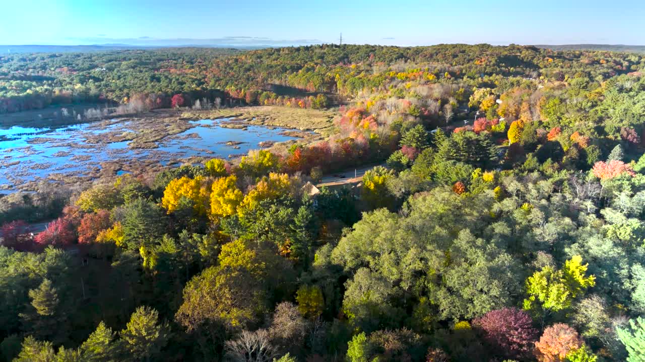 A breathtaking drone shot capturing a stunning sunset over a Massachusetts lake in autumn. Warm colors of the setting sun blend with the vibrant fall foliage, creating a serene and picturesque scene.