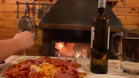 hand reaching out to pick up wine glass beside rustic pizza with fireplace in background