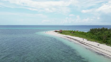 Tropical-beach-bungalow-huts-and-tourists-roam-white-sandy-beach-shoreline,-aerial