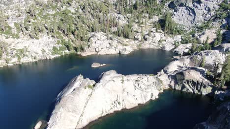 A-rock-island-sits-in-the-middle-of-a-clear-water-alpine-lake-at-the-base-of-a-mountain-range-in-the-wilderness-of-California-with-cliffs-and-people-camping-and-hiking-at-sunset-in-the-summer