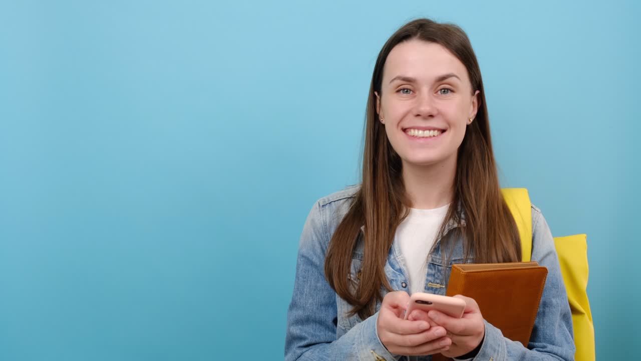 retrato de una niña sonriente estudiante adolescente usar chaqueta de denim y mochila amarilla sostener libros usar teléfono celular móvil mirar a un lado en el área de trabajo copiar el espacio maquillaje, aislado en la pared de fondo del estudio azul