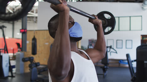 fit african american man wearing face mask exercising using barbell in the gym