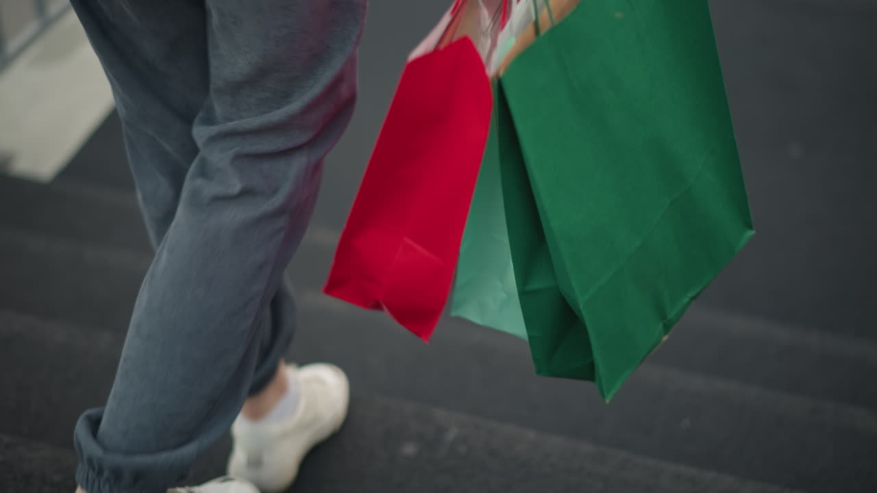 vista lateral de cerca de una mujer con ropa gris caminando por las escaleras mientras lleva bolsas de compras, está descendiendo lentamente en una alfombra negra, con una barandilla de hierro visible en el fondo borroso