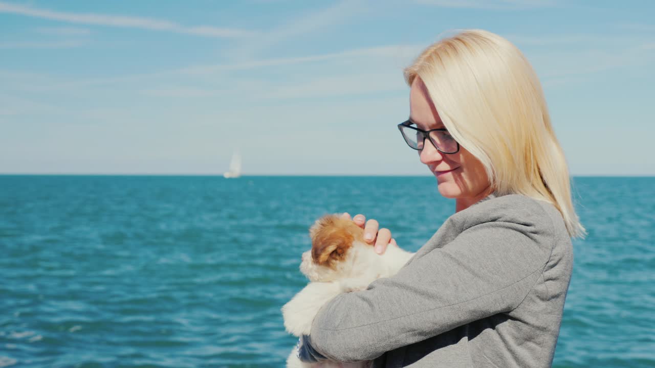 한 여인이 바다를 배경으로 강아지를 품에 안고 있다. 멀리서 요트를 볼 수 있다.