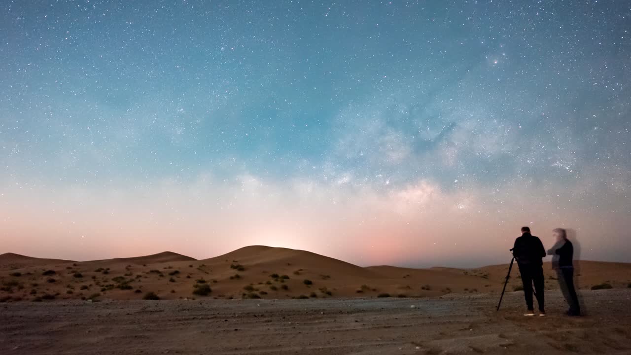 Two men setting up a panorama star tracker with the Milky Way rising almost parallel to the horizon in the desert sand dunes time lapse
