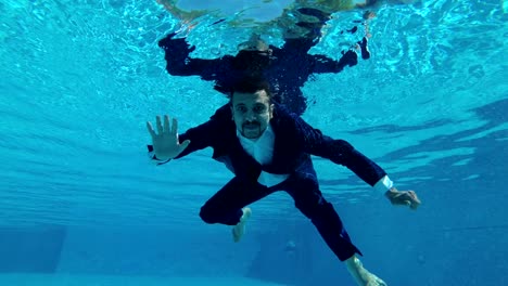 the groom in a suit swims and poses under the water in the pool. he looks at the camera, waves his hands and smiles. slow motion. action camera underwater.