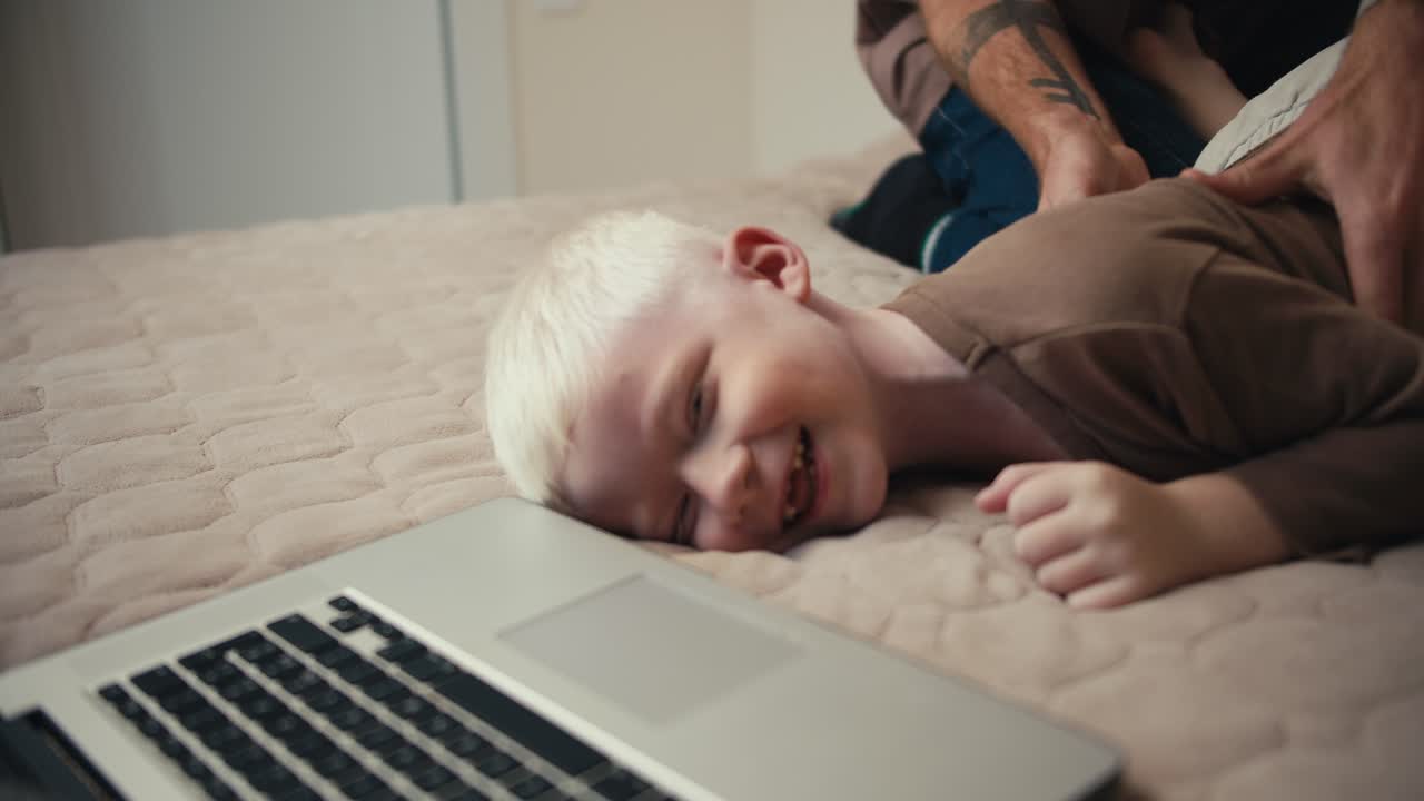 Close-up shot of a small albino boy with white hair in a brown sweater lying on the bed while his father tickles him lying near the laptop. Happy fun time for parents and children during vacation and weekend