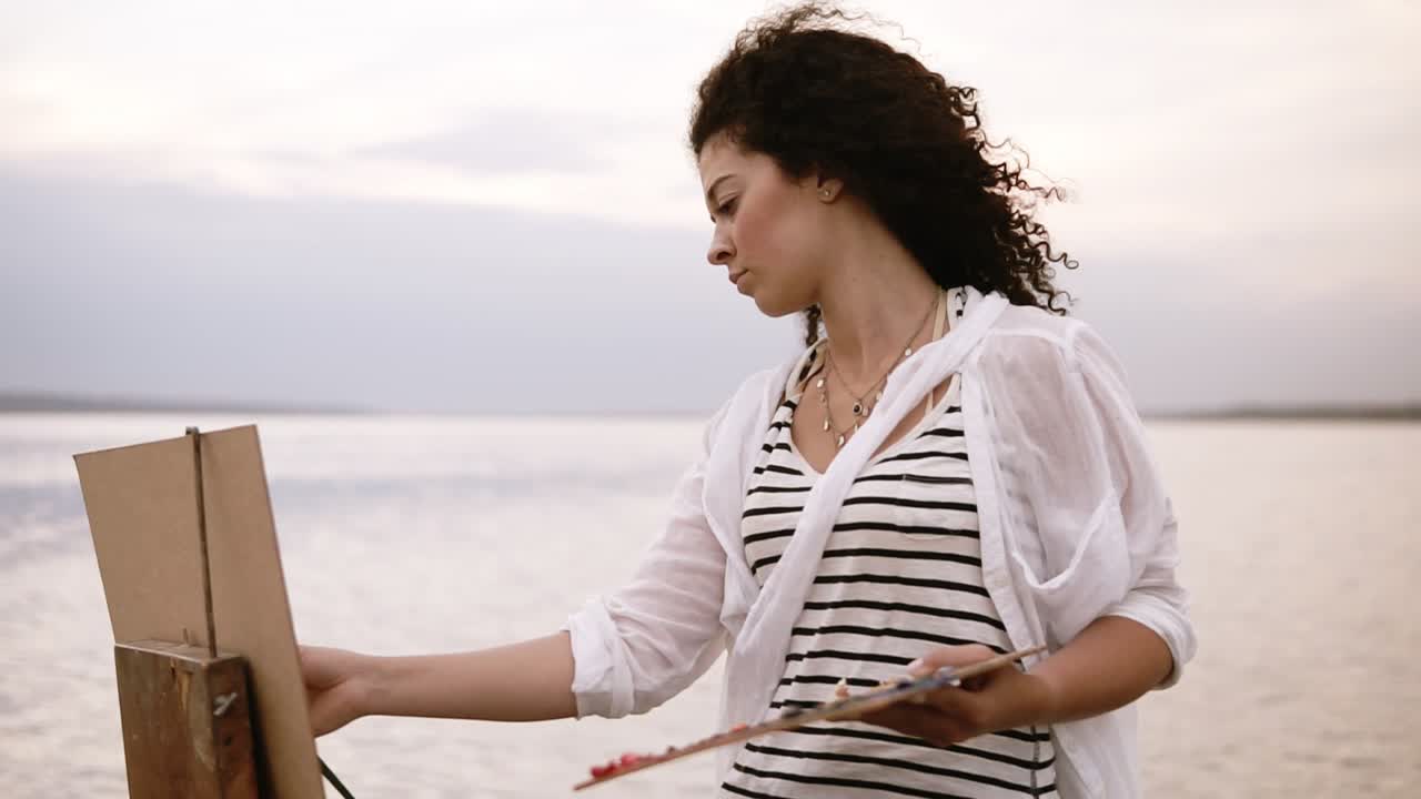 Close up of a gorgeous woman with brunette curly hair is standing in front the lake and drawing using a palette and an easel. Serious and concentrated. Morning clear sky