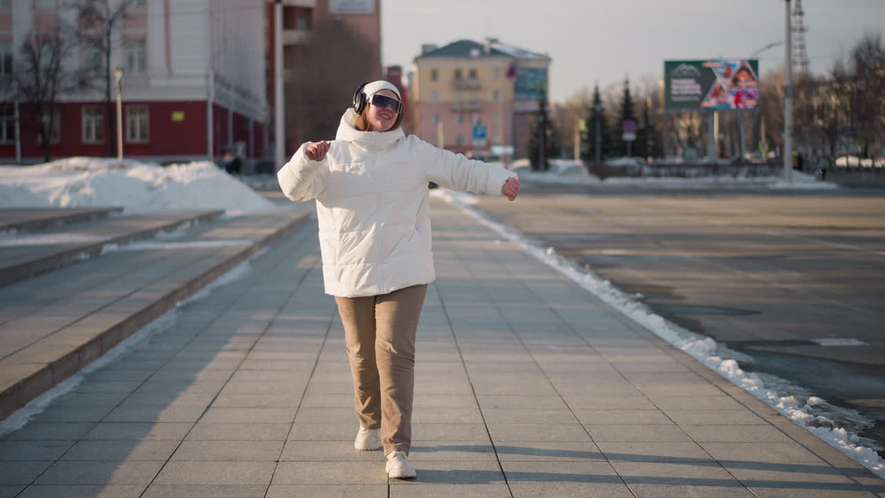 Young woman walking down tiled pavement in snowy urban park swaying arms to music wearing puffy white coat beanie sunglasses headphones playful dance moves and carefree winter stroll through city