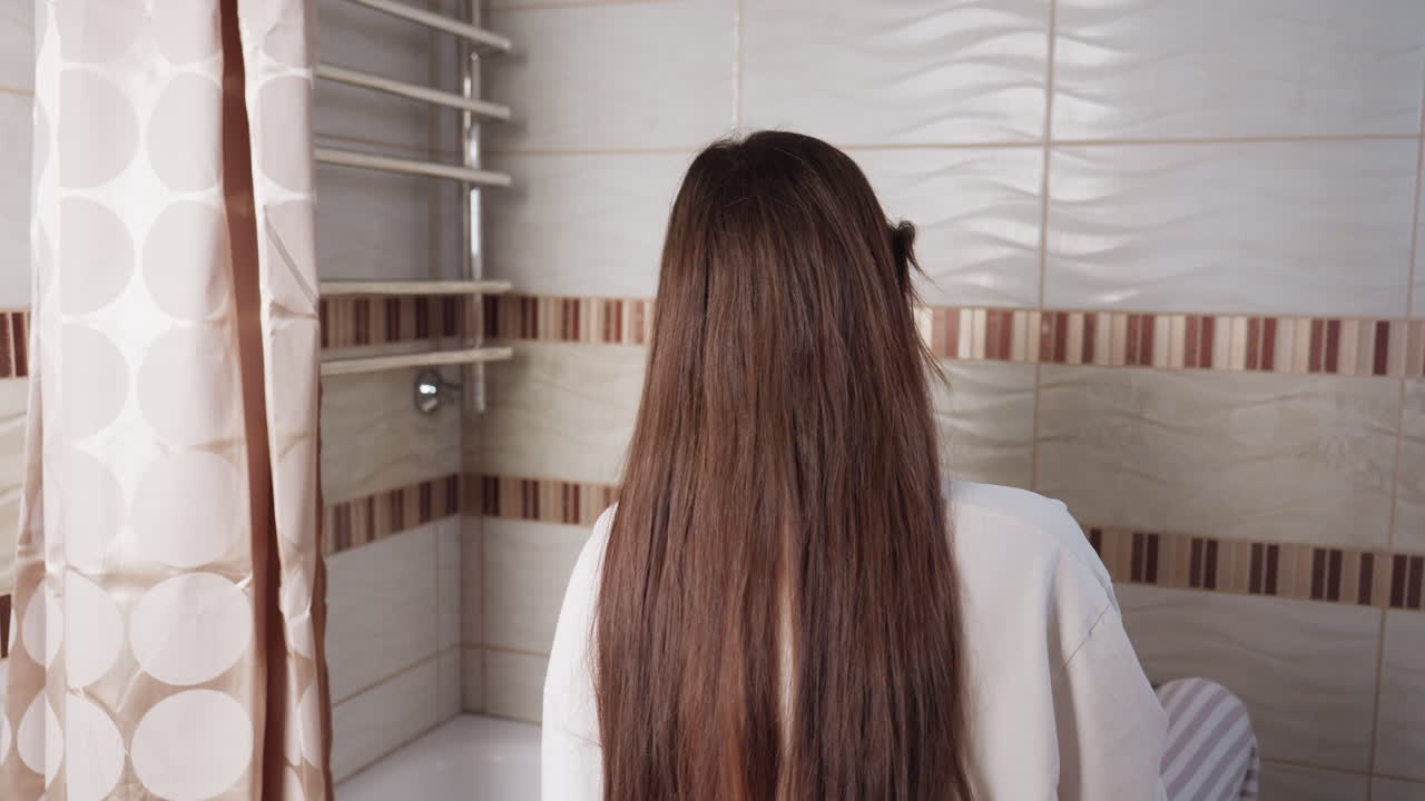 Back view student with long hair in tiled bathroom seated on bathtub reaches for towel on hanger, removing fabric from chrome rail beside curtain, everyday hygiene moment after wash