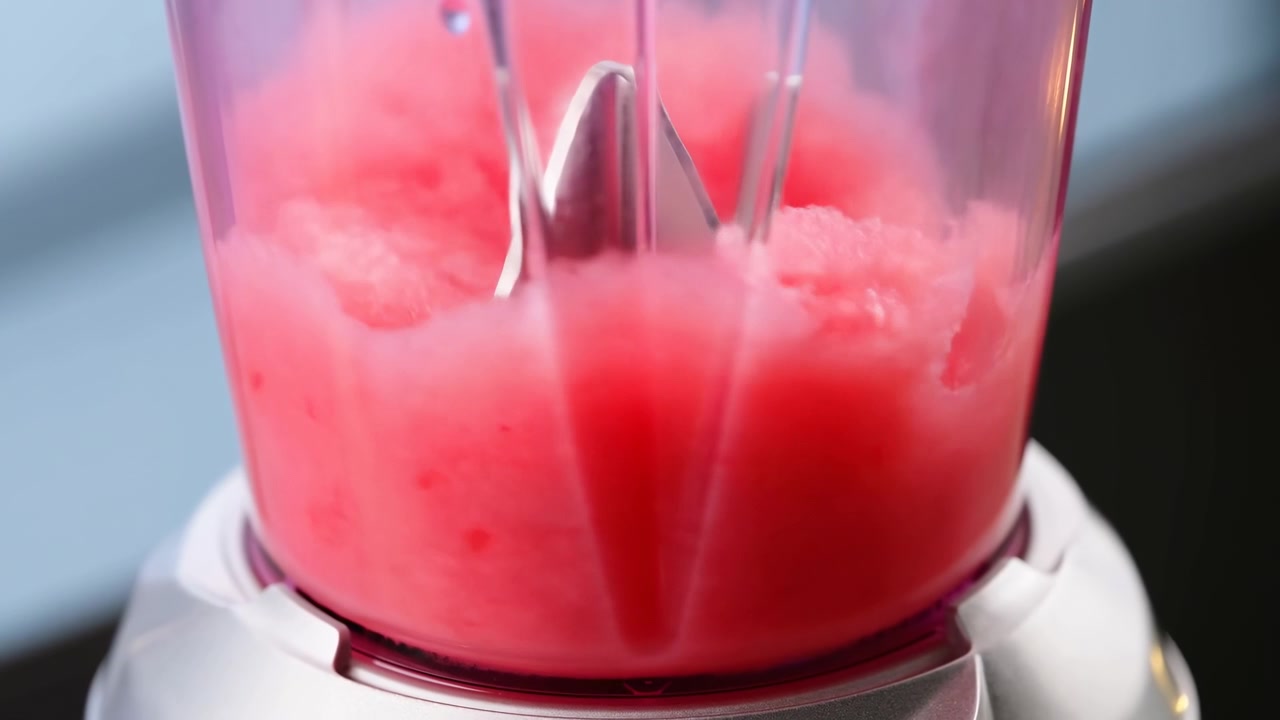 Primo piano all'altezza degli occhi di un boccale di frullatore riempito con una bevanda rosa, mentre il liquido vortica intorno alle lame, formando una texture liscia semi-opaca con morbidi riflessi, sottili striature e un'atmosfera da cucina moderna e pulita
