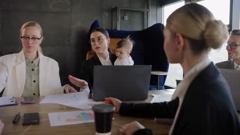 Zoom-in-brunette-girl-Business-Woman-with-glasses-in-business-clothes-holds-her-small-infant-daughter-in-her-arms-while-sitting-at-the-table-during-a-meeting-in-a-modern-office