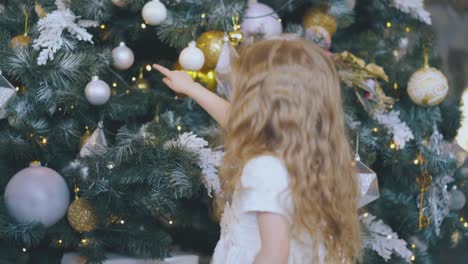 little-girl-in-dress-touches-balls-on-Christmas-tree-at-home