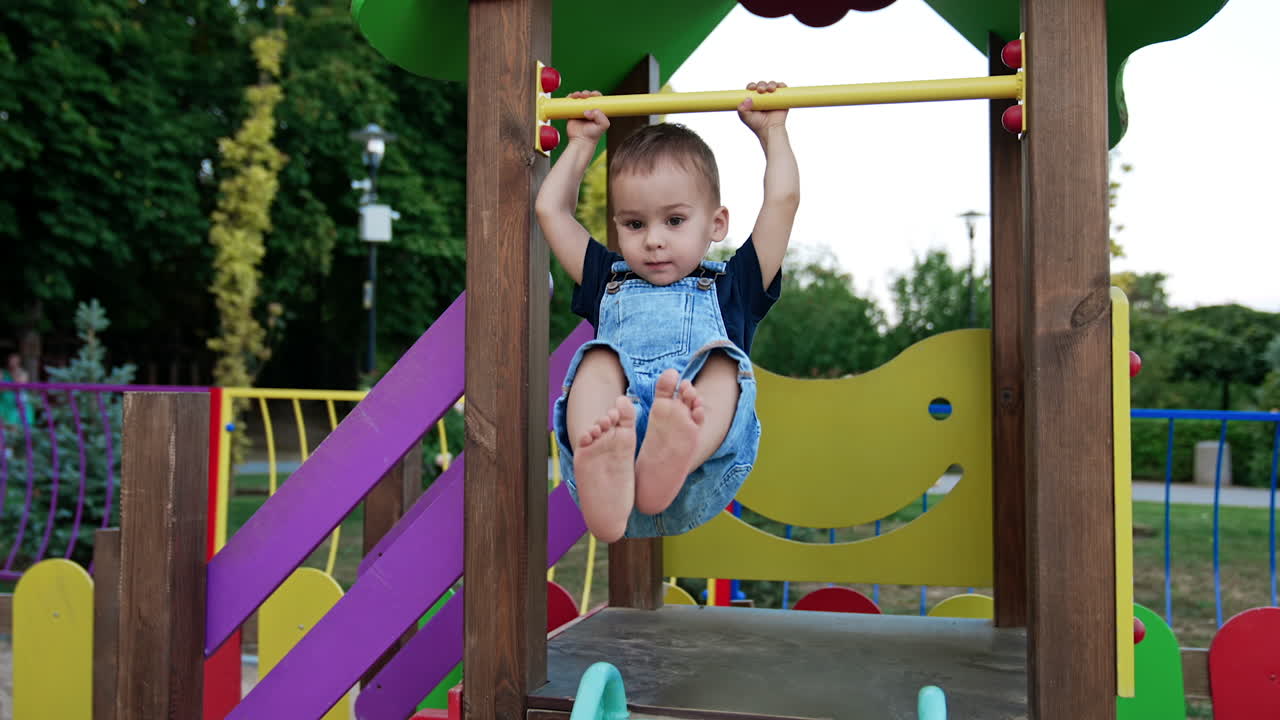 Adorable barefoot baby boy hanging on the horizontal bar. Smiling kid sits on the slide and hits his feet by it. Child on the playground in summer.