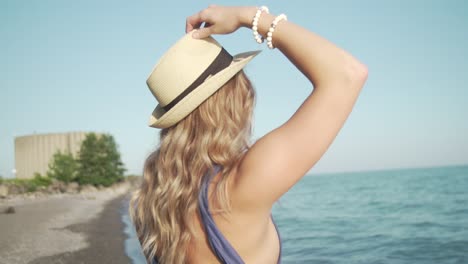 a blonde girl adjusts her fedora on the beach while looking at the water