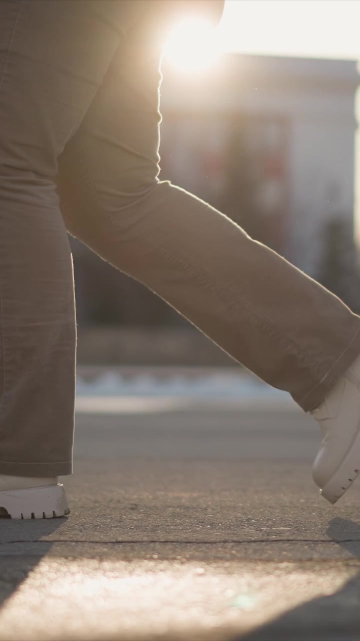 Person walking focusing on legs and white shoes with beige pants soft golden light creates warm shadows as person moves across pavement in outdoor setting