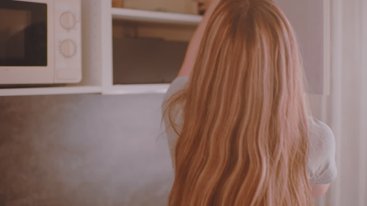 Natural light streams into kitchen as back view of woman in grey short pants with long flowing hair walks toward cabinet to collect item, creating casual morning household routine