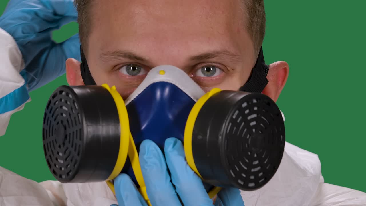 портрет мужского врача в белом защитном костюме и хирургических перчатках, глядящий в камеру, надевает медицинский респиратор. зеленый экран, хроматический ключ. замедленное движение