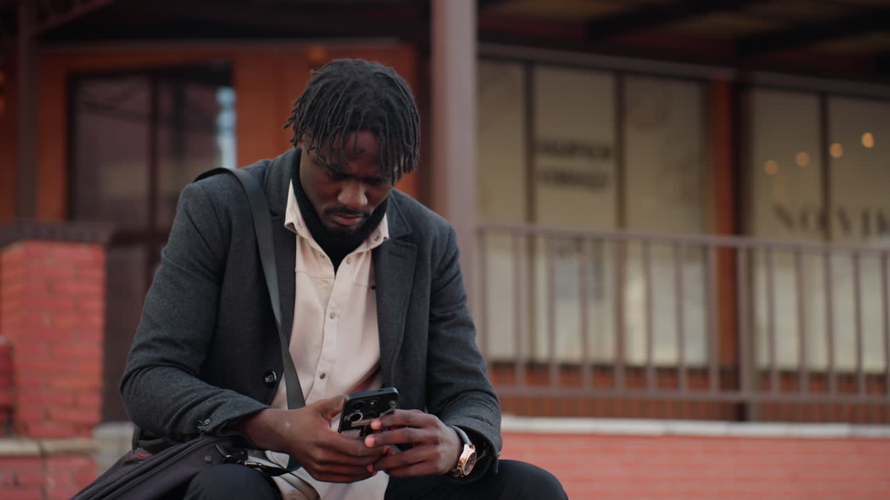Campus student wearing layered outfit sits outdoors concentrating on phone screen with serious facial expression, showing focus, determination, and digital engagement in calm urban environment