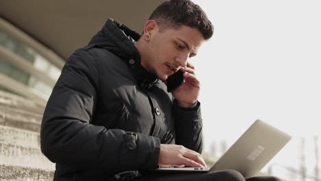 Focused-young-man-talking-on-phone-and-typing-on-laptop