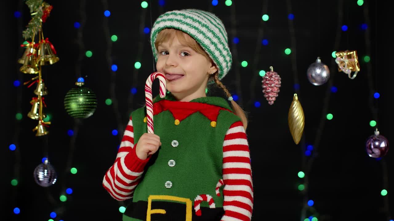Kid girl in Christmas elf Santa Claus helper costume licking candy cane lollipop caramel sweets