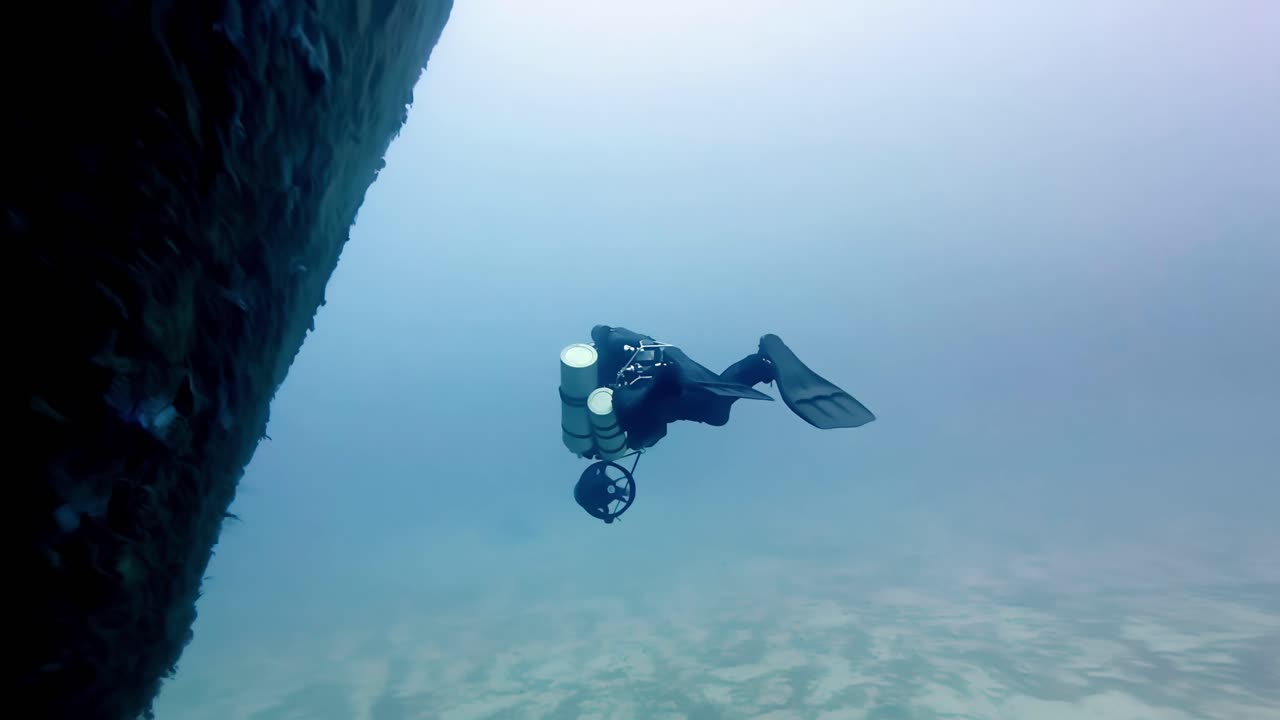Esploratore subacqueo naviga nelle profondità blu vicino a formazioni rocciose sottomarine aspre a Malta, Gozo e Comino, durante una limpida giornata estiva, catturato tramite uno scatto grandangolare dinamico che mostra vivide ombre marine.
