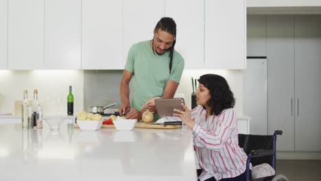 Happy-biracial-woman-in-wheelchair-using-tablet,-preparing-food-with-male-partner-in-kitchen