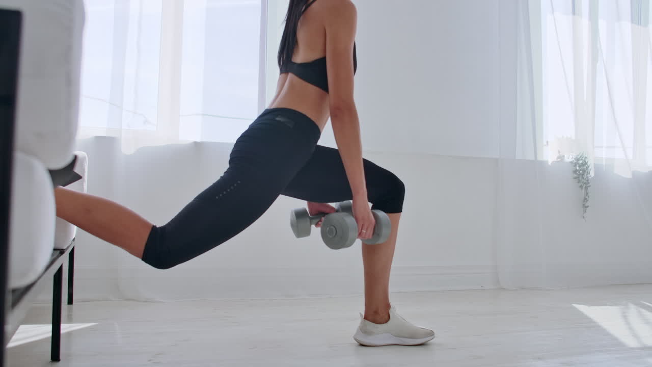 Una bruna in abbigliamento sportivo nero in un appartamento bianco esegue uno split squat con manubri tra le mani, appoggiando il piede sul divano.