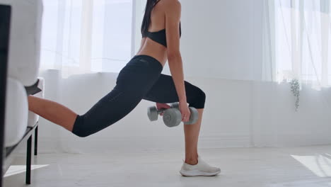 brunette in black sportswear in a white apartment makes a split squat with dumbbells in her hands leaning on the sofa with her foot.