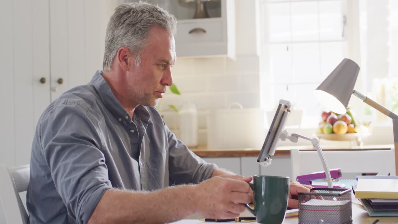 Happy caucasian man sitting at table in kitchen, using tablet and drinking coffee
