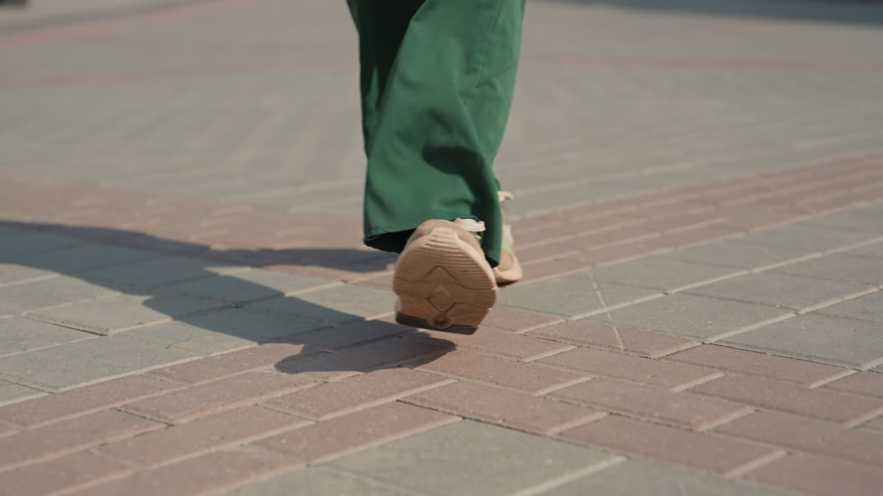 Closeup walking sneakers on sunlit pavement, green wideleg trousers sway with each confident step through textured brick plaza, soft shadows cast by midday sun, casual urban commute mood, focus