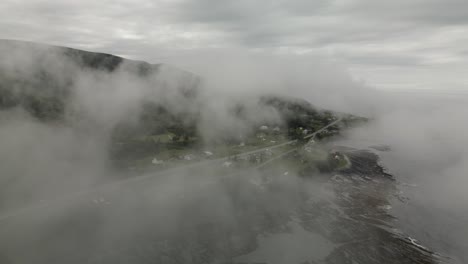 looking through gloomy fog over the quaint green coastal chic-chocs village, drone orbit with tilt down